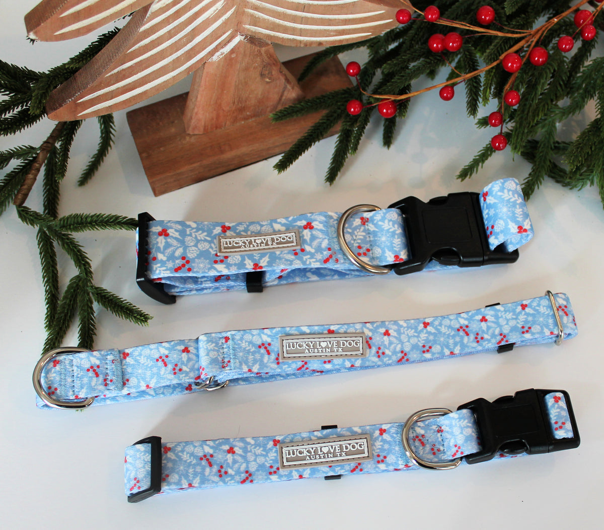 Light blue winter-themed Lucky Love Dog collars with snowflake and berry patterns displayed alongside festive holiday décor.
