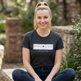 A smiling woman with blonde hair in a ponytail sits cross-legged outdoors wearing a dark charcoal heather t-shirt with a white rectangular chest graphic that reads 'LUCKY LOVE DOG AUSTIN TX' and 'FOR THE LOVE OF DOGS'.