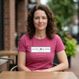 A woman with curly brown hair sits at an outdoor cafe table wearing a pink t-shirt with a white graphic that reads 'LUCKY LOVE DOG AUSTIN, TX'.