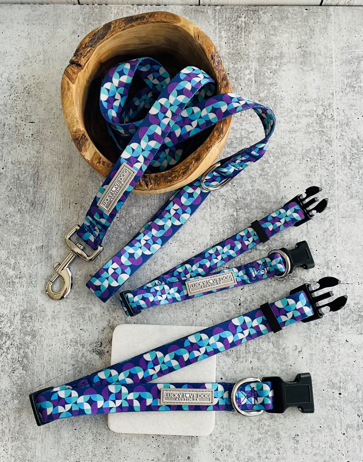 Set of Lucky Love Dog Jasper collection accessories displayed on a stone surface, including a matching geometric blue collar, leash, and waste bag holder in a wooden bowl.