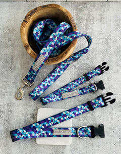 Set of Lucky Love Dog Jasper collection accessories displayed on a stone surface, including a matching geometric blue collar, leash, and waste bag holder in a wooden bowl.